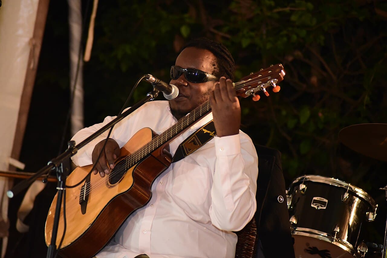 A visually impaired Kenyan musician singing into a microphone and playing an acoustic guitar